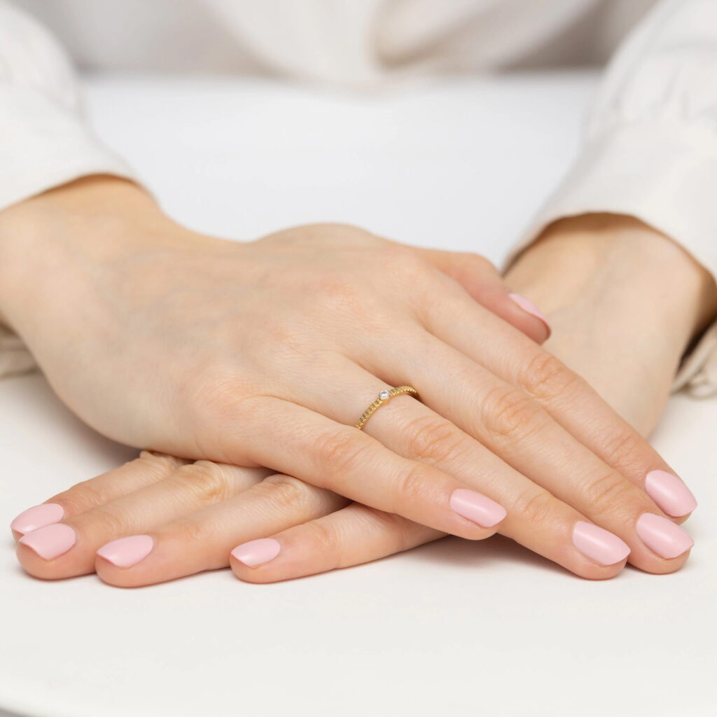 Dwie dłonie z różowym manicure, jedna z cienką złotą obrączką z diamentem, ułożone na białej powierzchni.