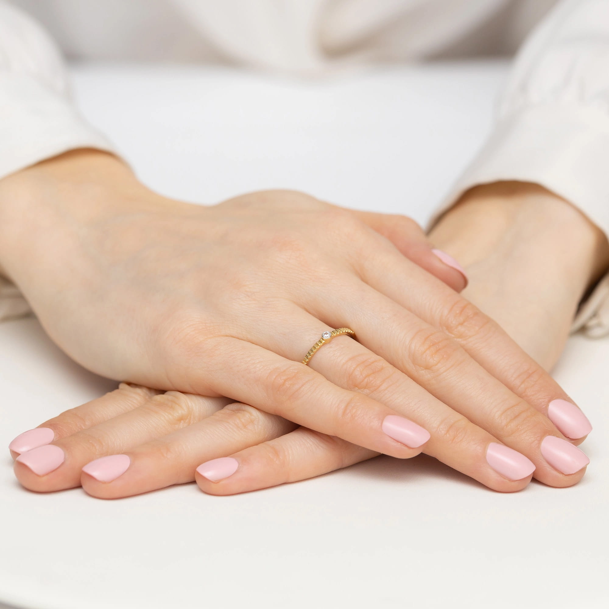 W0512.Z3.DIW.01_4 Dwie dłonie z różowym manicure, jedna z cienką złotą obrączką z diamentem, ułożone na białej powierzchni.