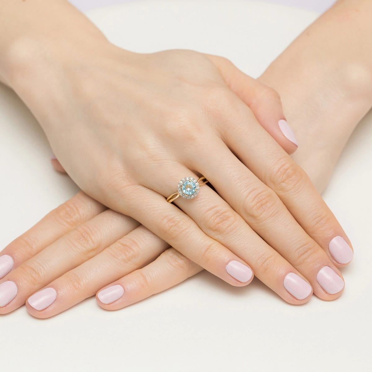 W0538.Z3.TOB.01_4 Złoty pierścionek z niebieskim kamieniem na dłoniach z delikatnym manicure w jasnym kolorze.