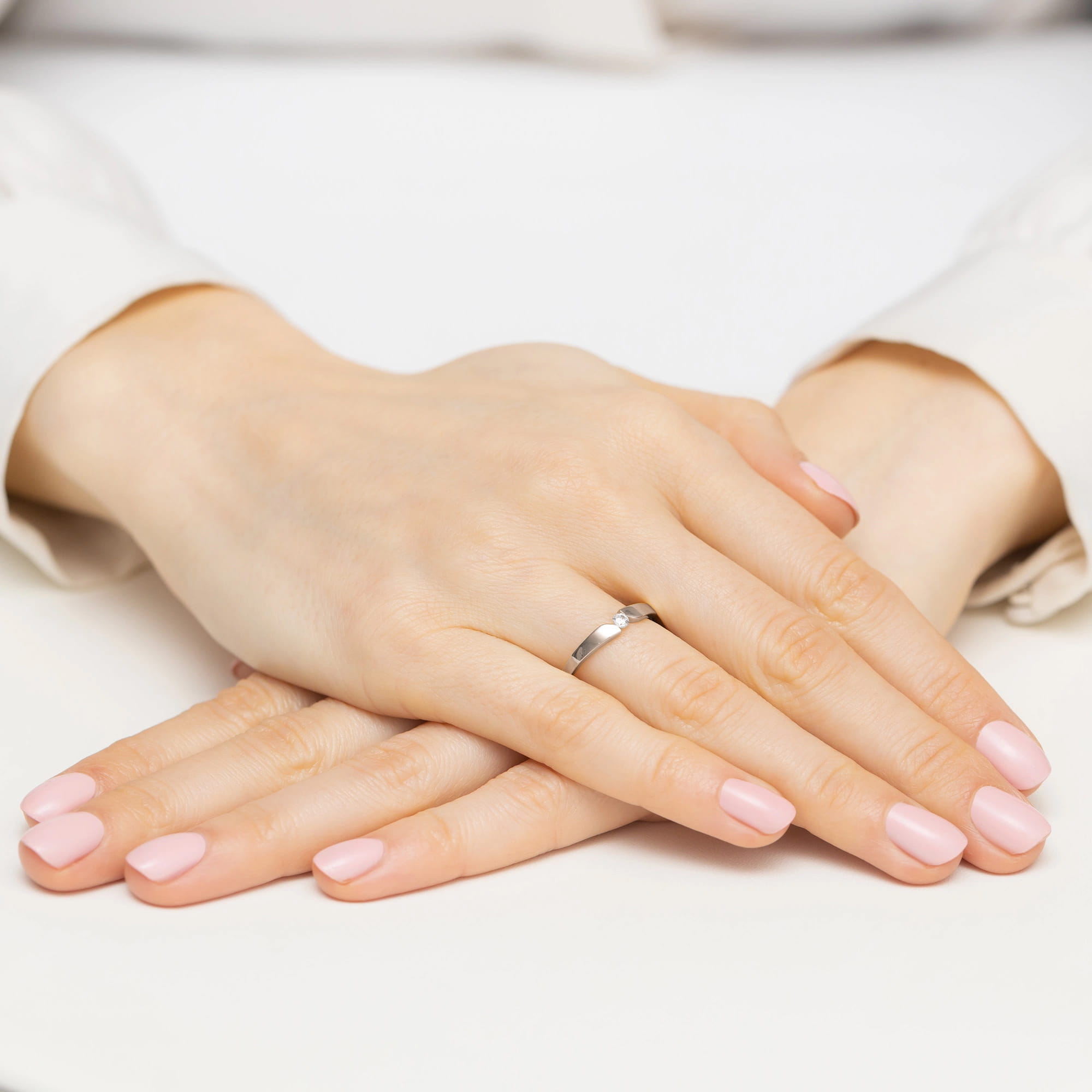 W0554.B3.DIW.01_4 Dwie ręce z delikatnym pierścionkiem i różowym manicure, ułożone na białym tle.