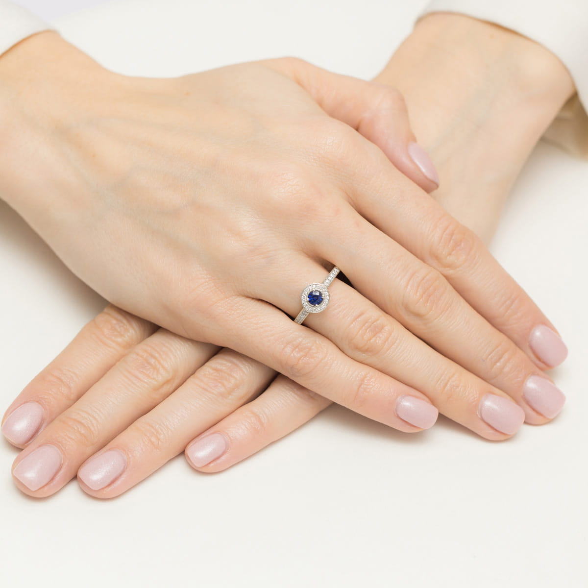 W0559.Z3.SAB.01_4 Dwie dłonie z delikatnym manicure, na jednej z nich pierścionek z niebieskim kamieniem i diamentami.