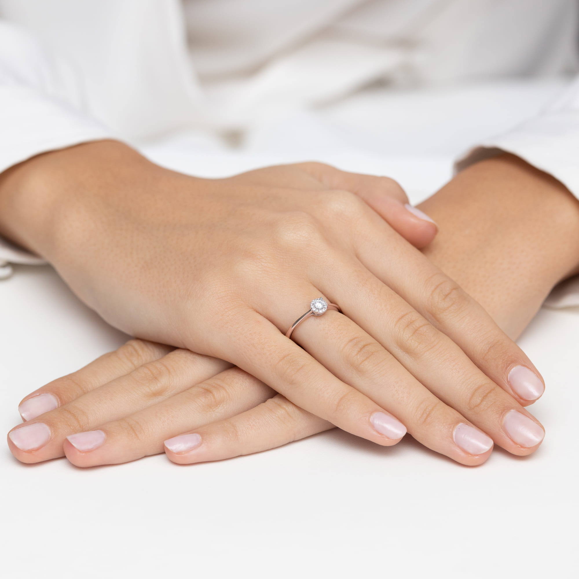 W0560.R3.DIW.01_4 Na dłoniach z delikatnym manicure znajduje się pierścionek z diamentem, ręce złożone na stole.