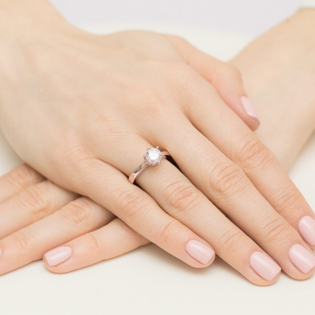 Na dłoni z delikatnym manicure znajduje się pierścionek z dużym, błyszczącym kamieniem.