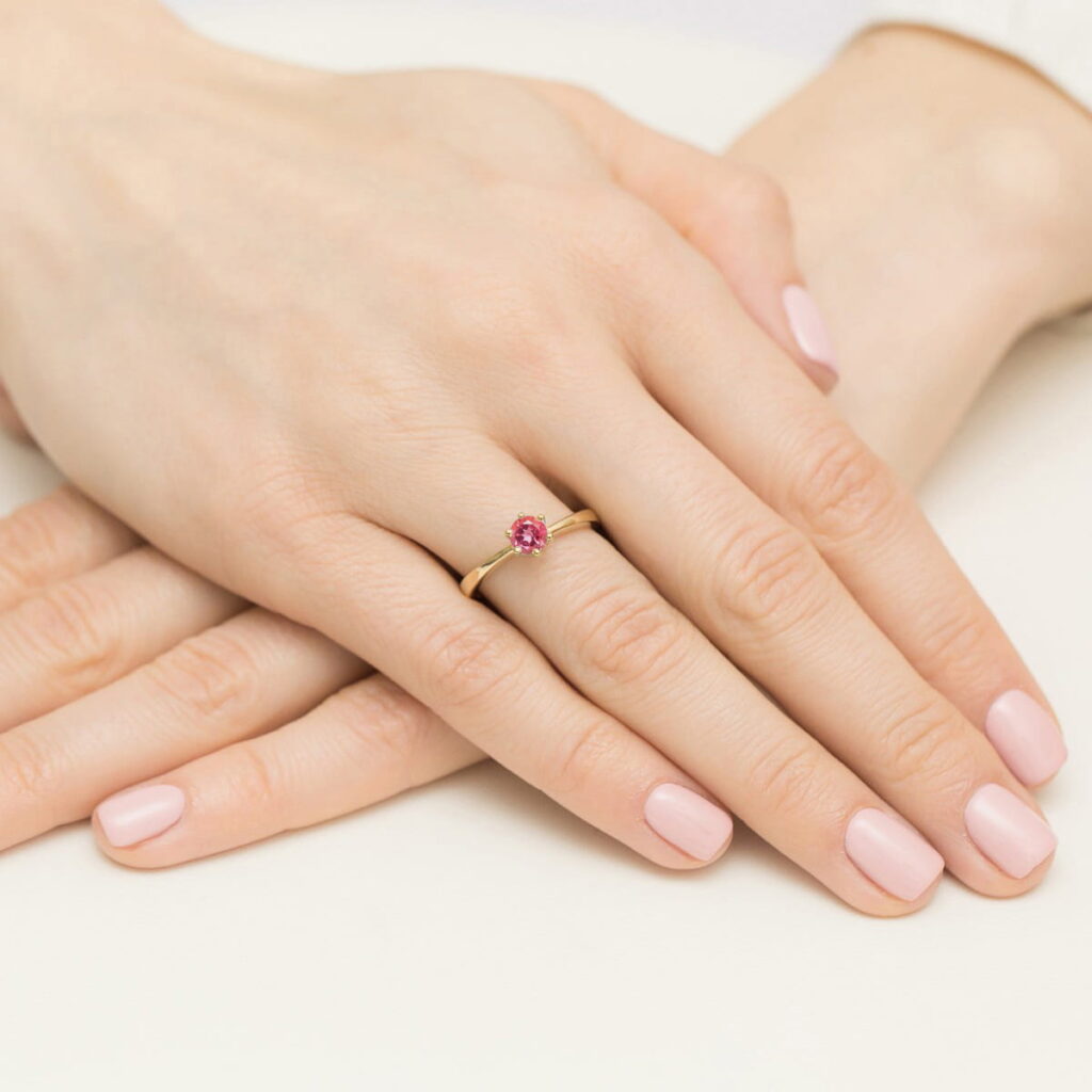 Złoty pierścionek z różowym kamieniem na palcu, dłonie z delikatnym manicure w jasnym kolorze.