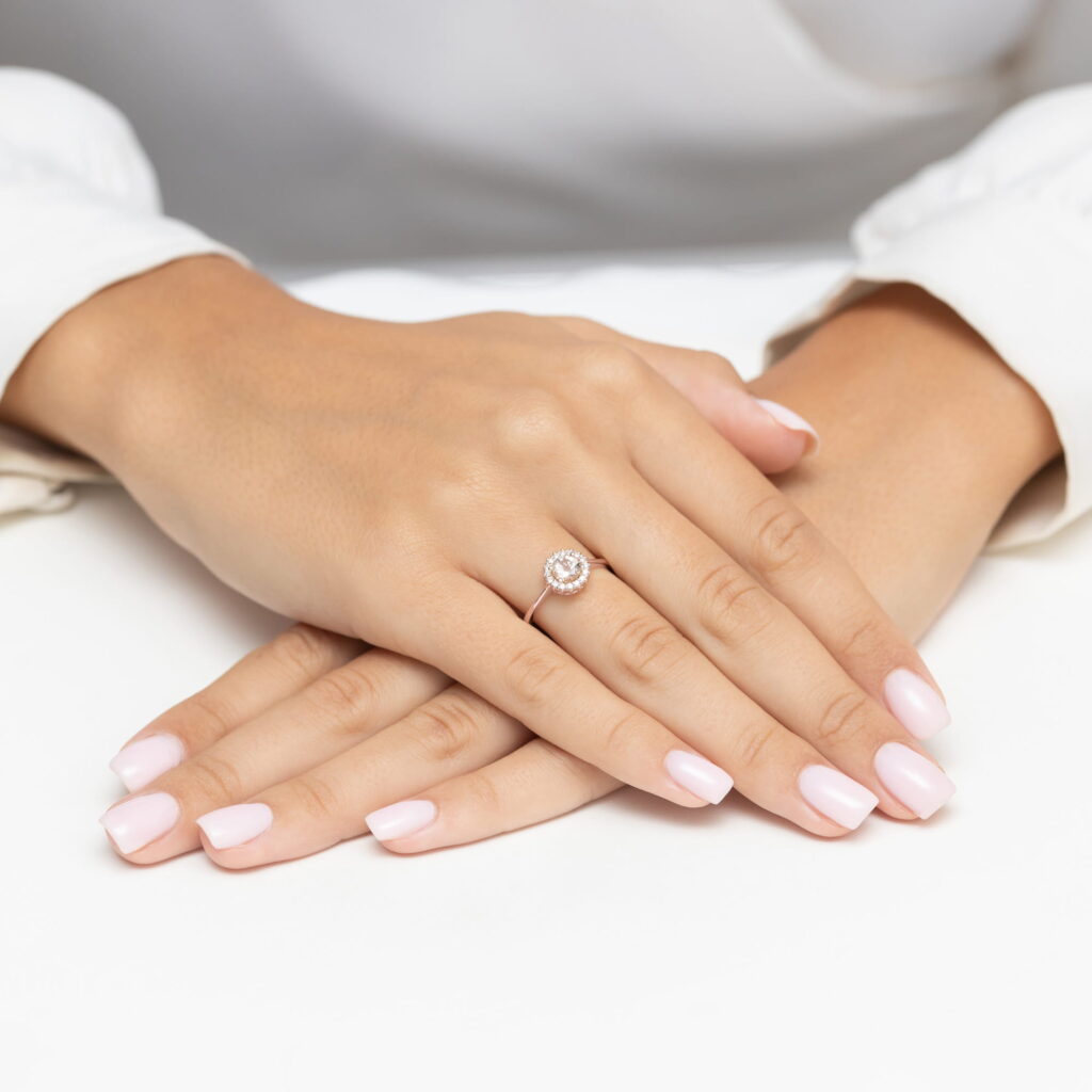 Dwie dłonie z różowym manicure, jedna z pierścionkiem z okrągłym kamieniem na palcu.