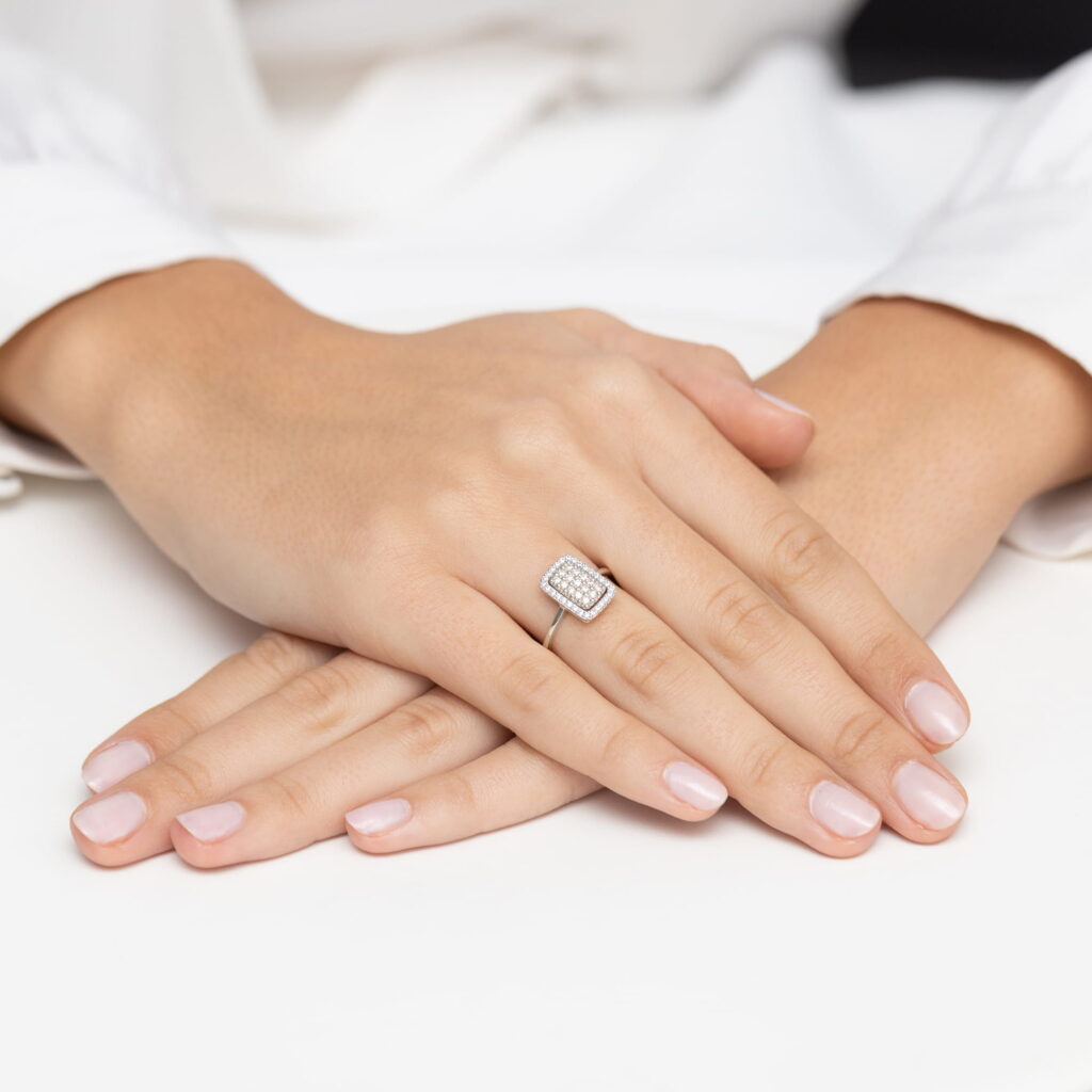 Dwie dłonie z delikatnym manicure, trzymające pierścionek z diamentami na palcu.