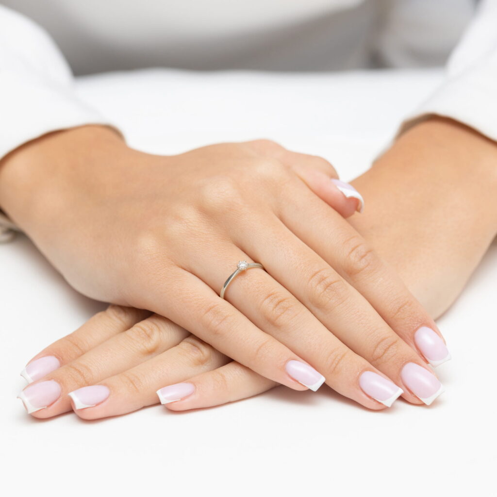 Dwie dłonie z delikatnym manicure, jedna z pierścionkiem, ułożone na białym tle.