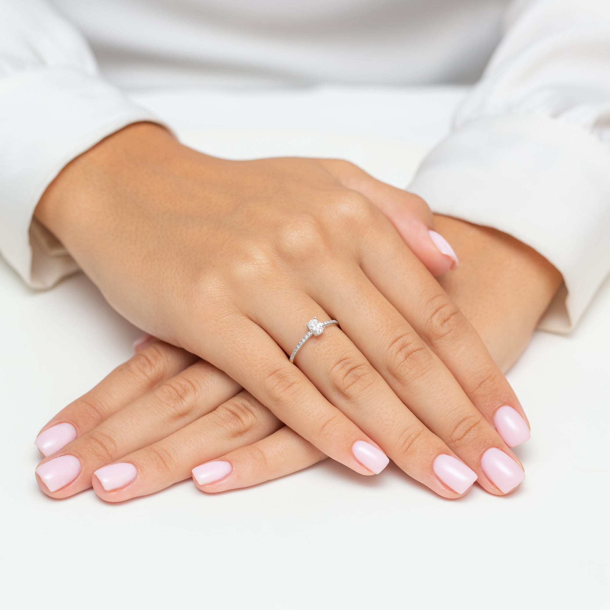 W0582.B3.DIW.01_4 Dwie dłonie z delikatnym pierścionkiem na palcu, z różowymi paznokciami, ułożone na białym tle.