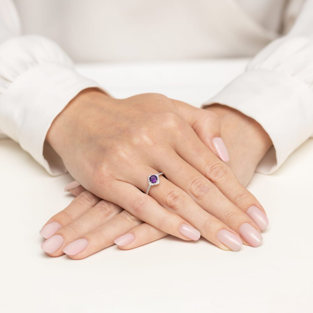 Na dłoni z delikatnym manicure znajduje się pierścionek z fioletowym kamieniem szlachetnym.