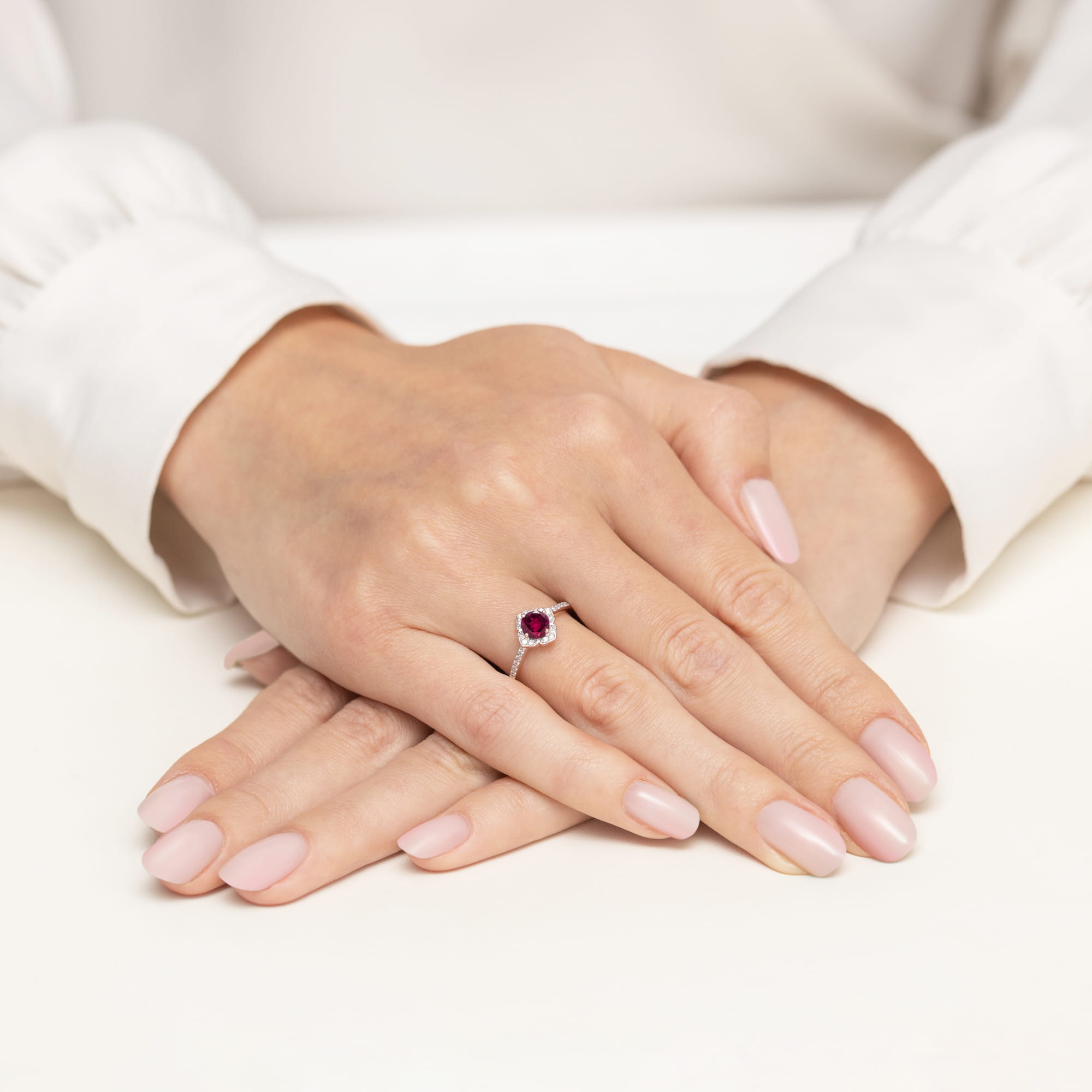 W0603.R3.RHR.01_4 Na dłoni widać pierścionek z rubinem, otoczony drobnymi diamentami, na tle jasnego stołu.