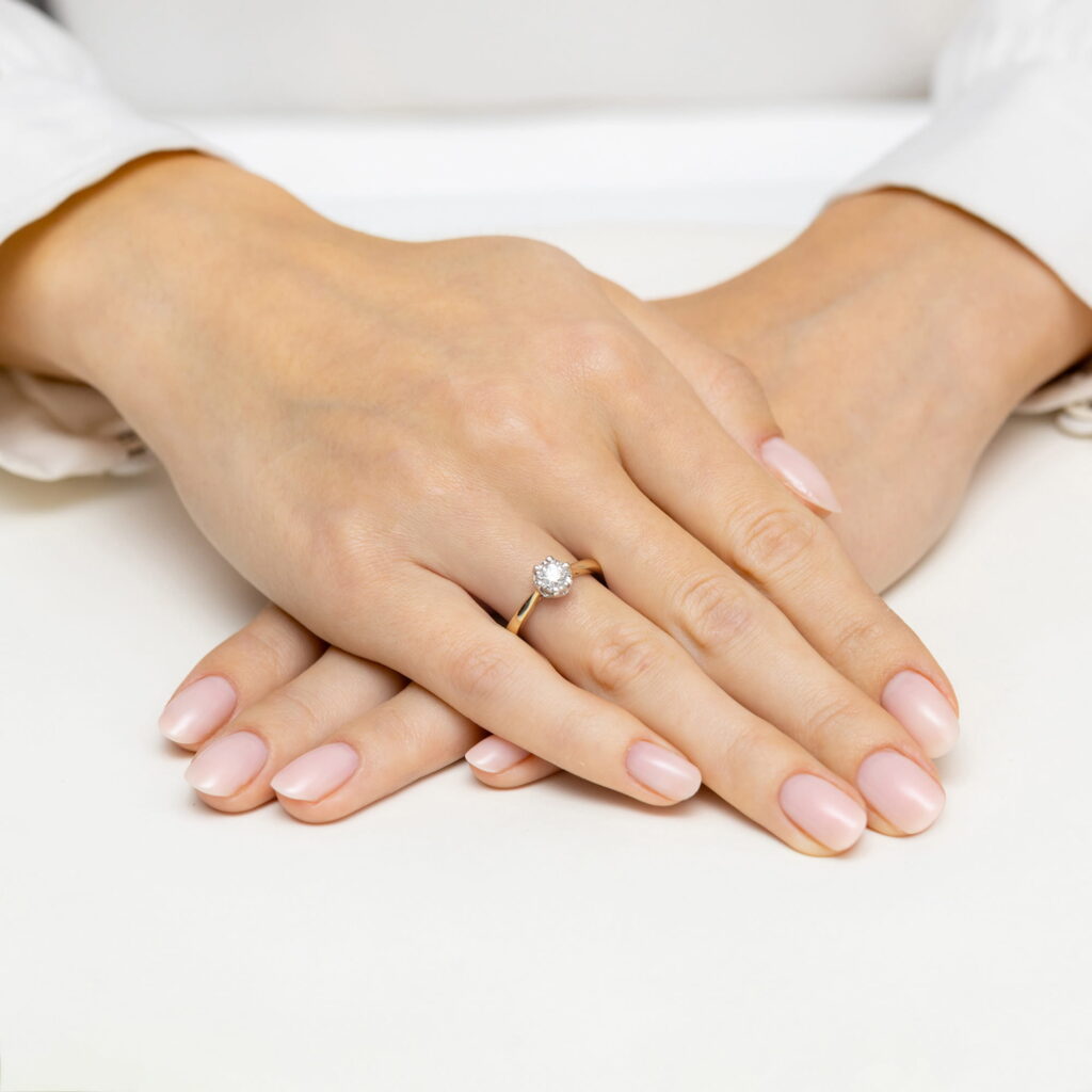 Dwie ręce z delikatnym manicure, jedna z pierścionkiem z dużym diamentem na złotej obrączce.
