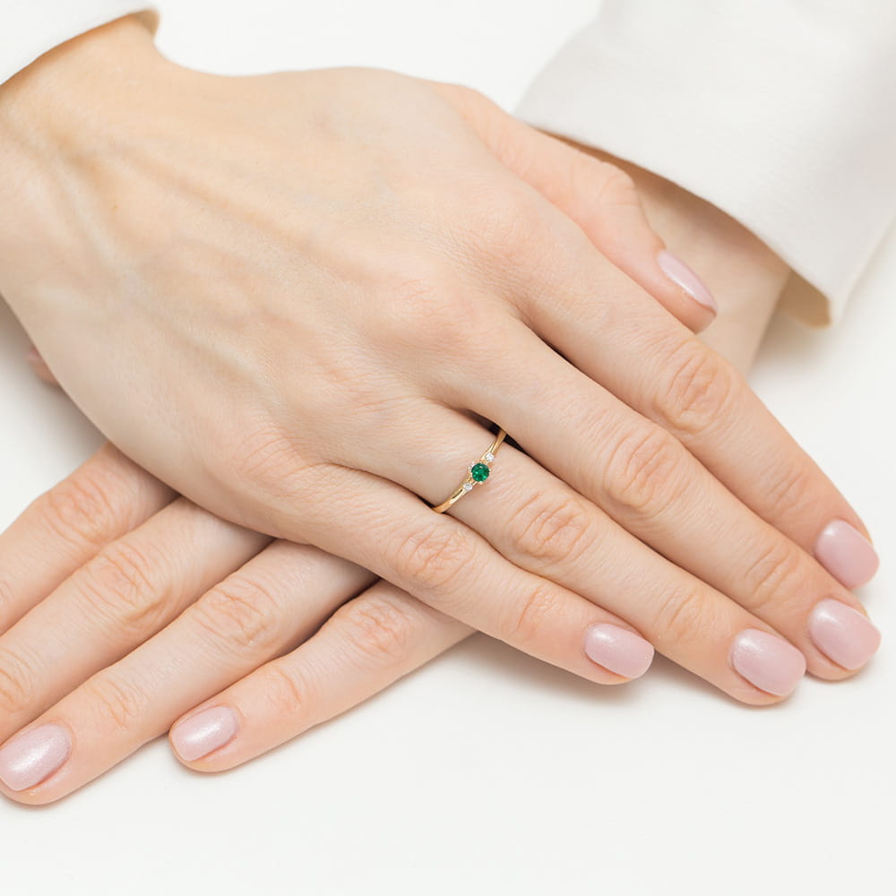 W0614.Z3.EMG.01_4 Dwie ręce z delikatnym pierścionkiem z zielonym kamieniem na palcu, paznokcie w jasnym kolorze.