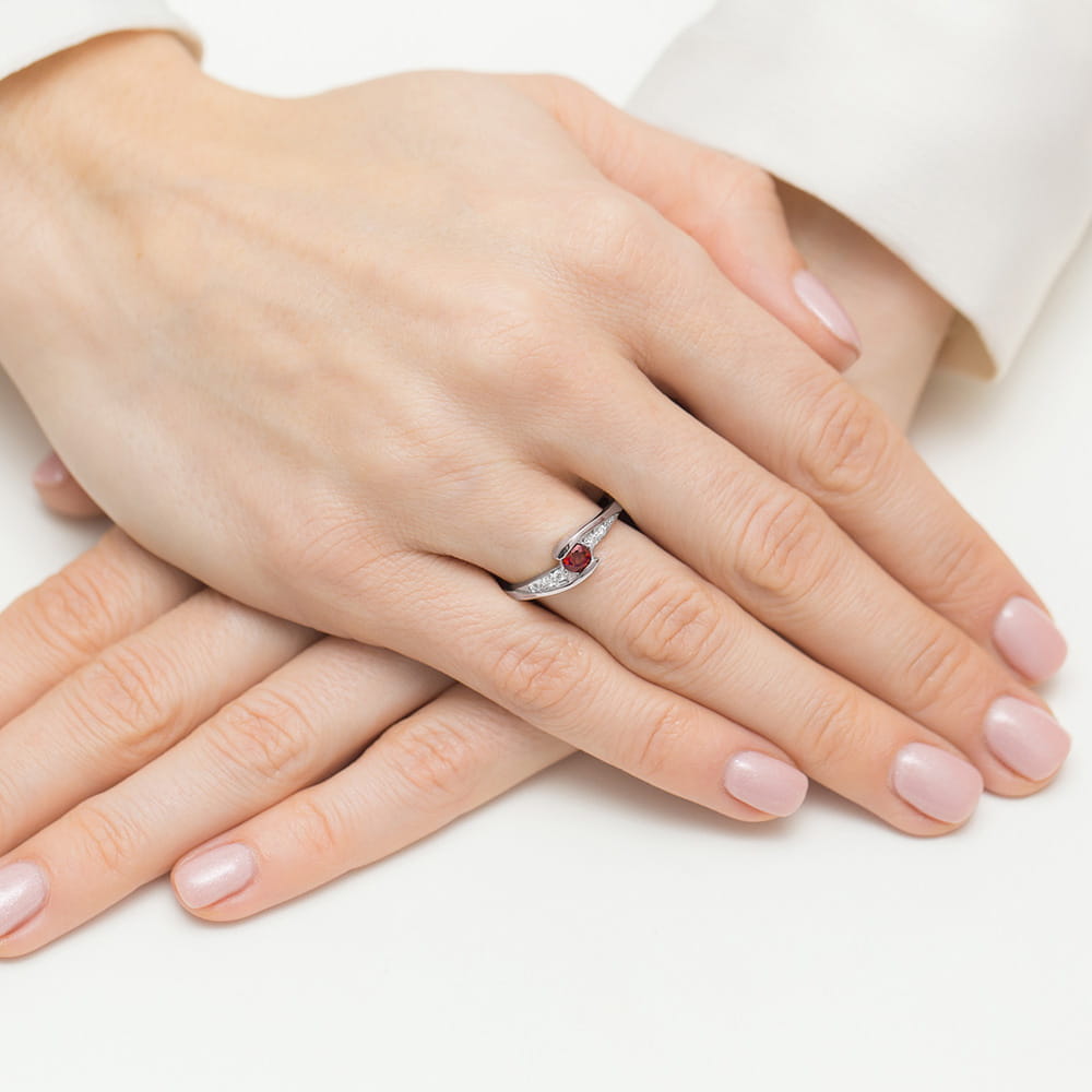W0618.B3.RUR.01_4 Na dłoni kobiety znajduje się srebrny pierścionek z czerwonym kamieniem, otoczony drobnymi diamentami.