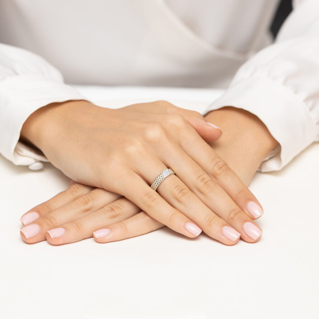 Dwie dłonie z delikatnym manicurem, jedna z pierścionkiem z diamentami, spoczywają na białym stole.