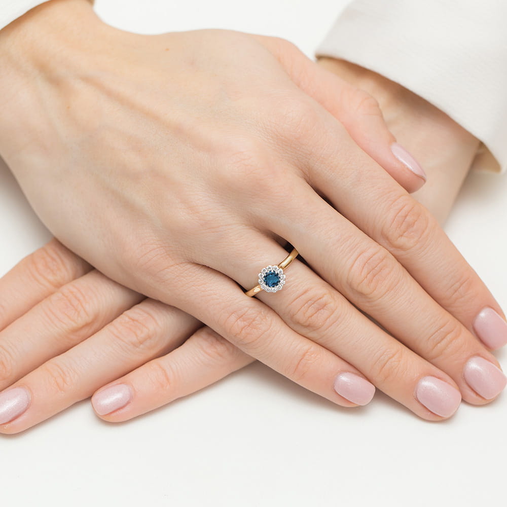 W0622.Z3.TOL.01_4 Złoty pierścionek z niebieskim kamieniem w otoczeniu drobnych diamentów na dłoni z różowymi paznokciami.