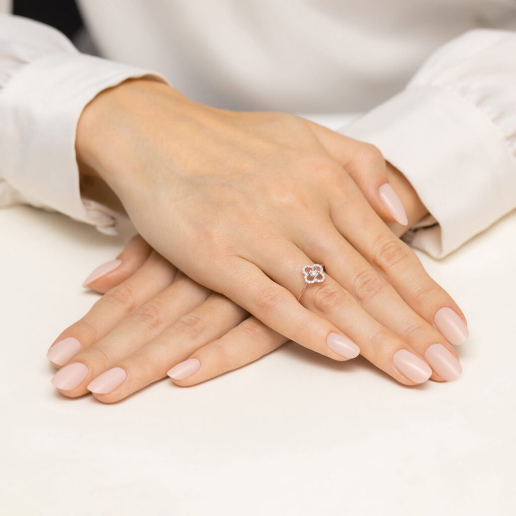 Dwie ręce z delikatnym manicure, jedna z pierścionkiem w kształcie kwiatu, spoczywają na jasnym tle.