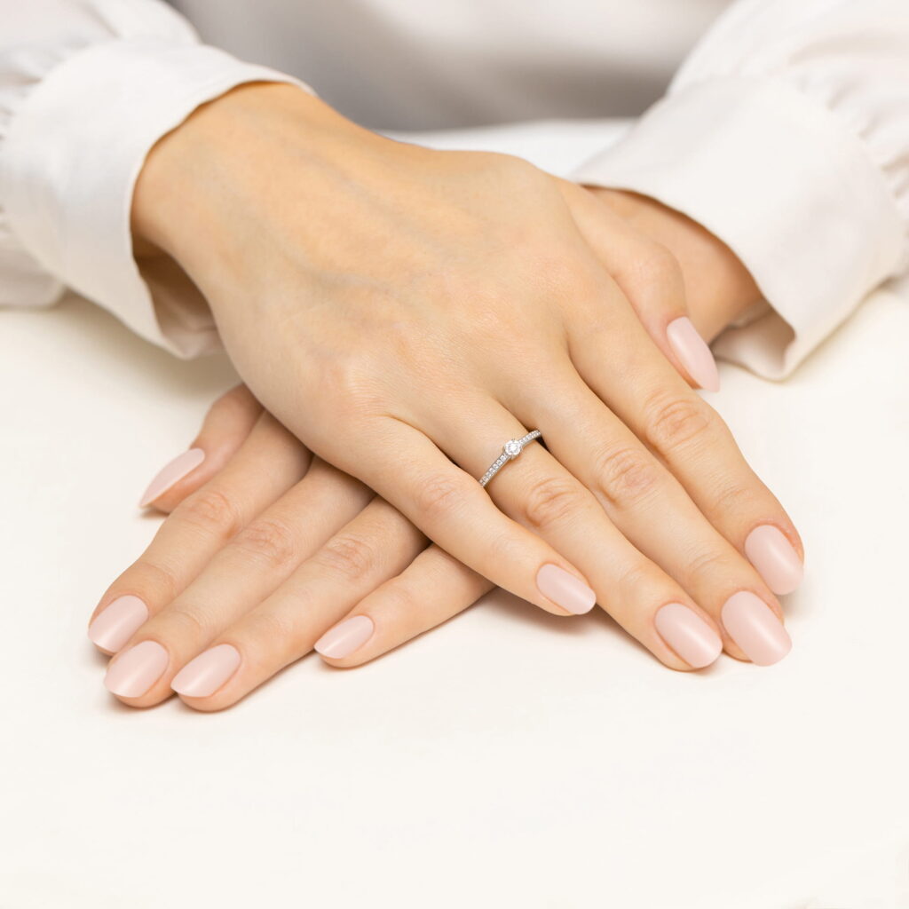 Dwie ręce z delikatnym manicure, jedna z pierścionkiem z diamentem, spoczywają na jasnym tle.