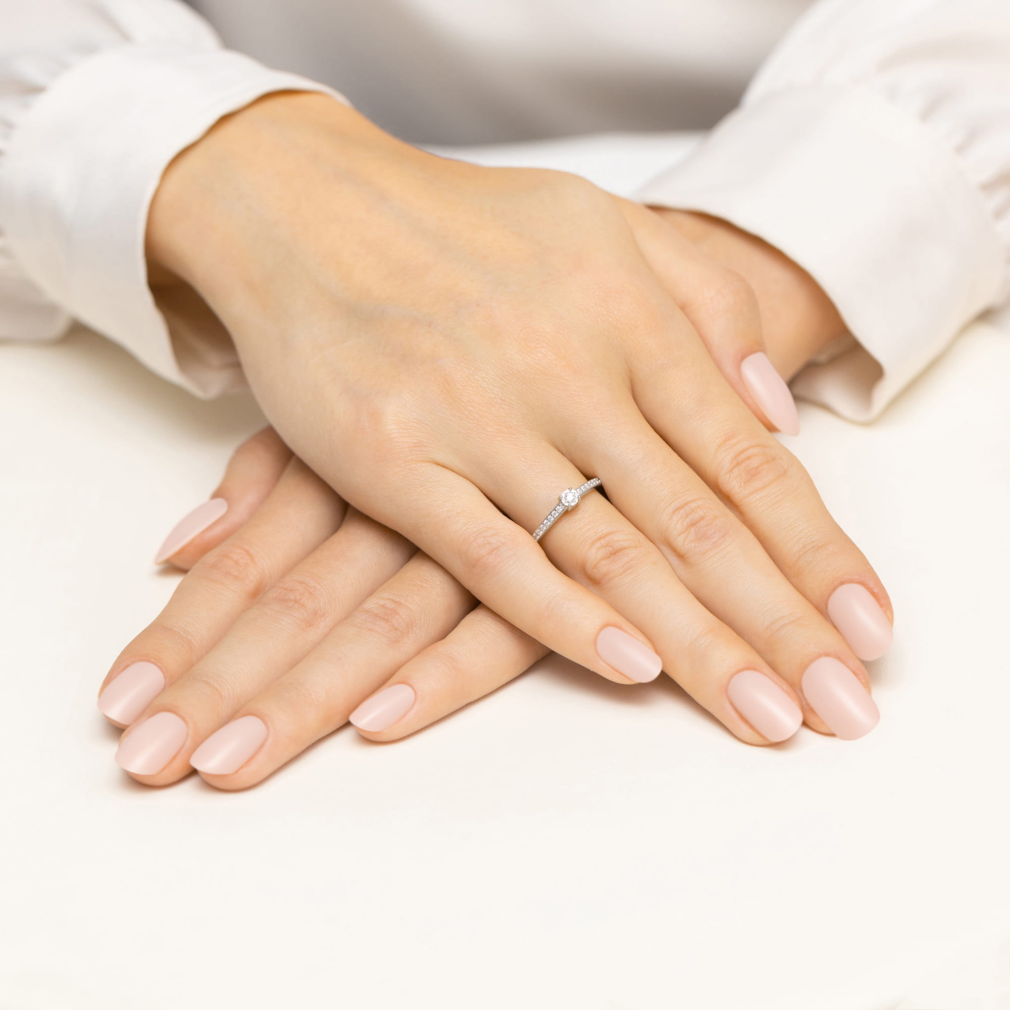 W0648.B3.DIW.01_4 Dwie ręce z delikatnym manicure, jedna z pierścionkiem z diamentem, spoczywają na jasnym tle.