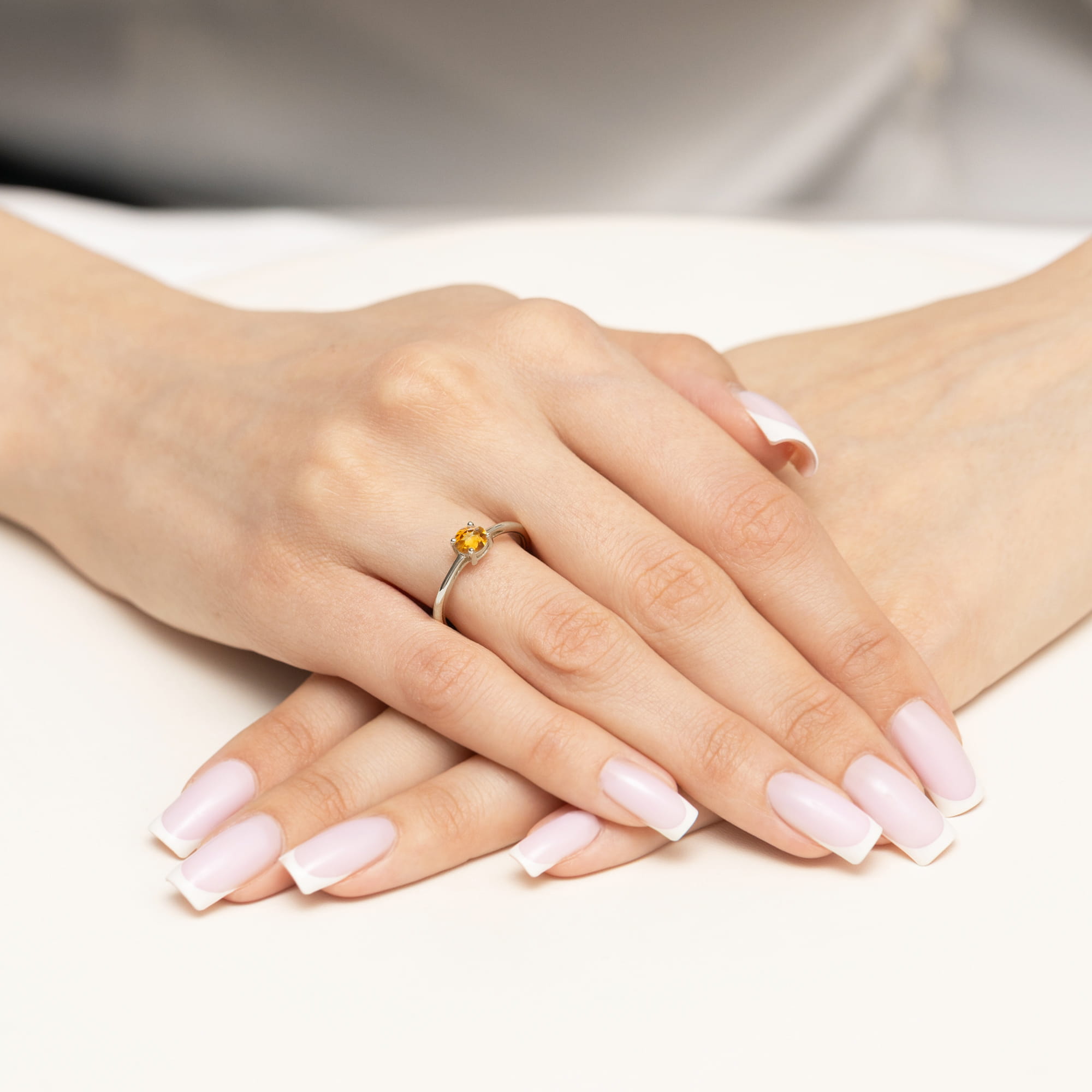 W0656.B3.CIY.01_4 Dłonie z długimi, jasnymi paznokciami trzymają pierścionek z żółtym kamieniem na białym tle.