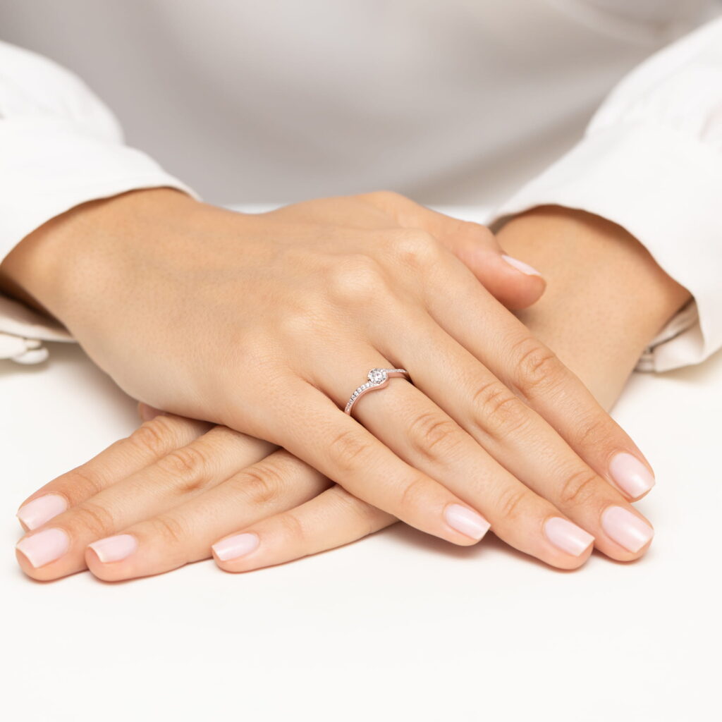 Dwie ręce z delikatnym manicure, jedna z pierścionkiem z diamentem, spoczywają na białym tle.