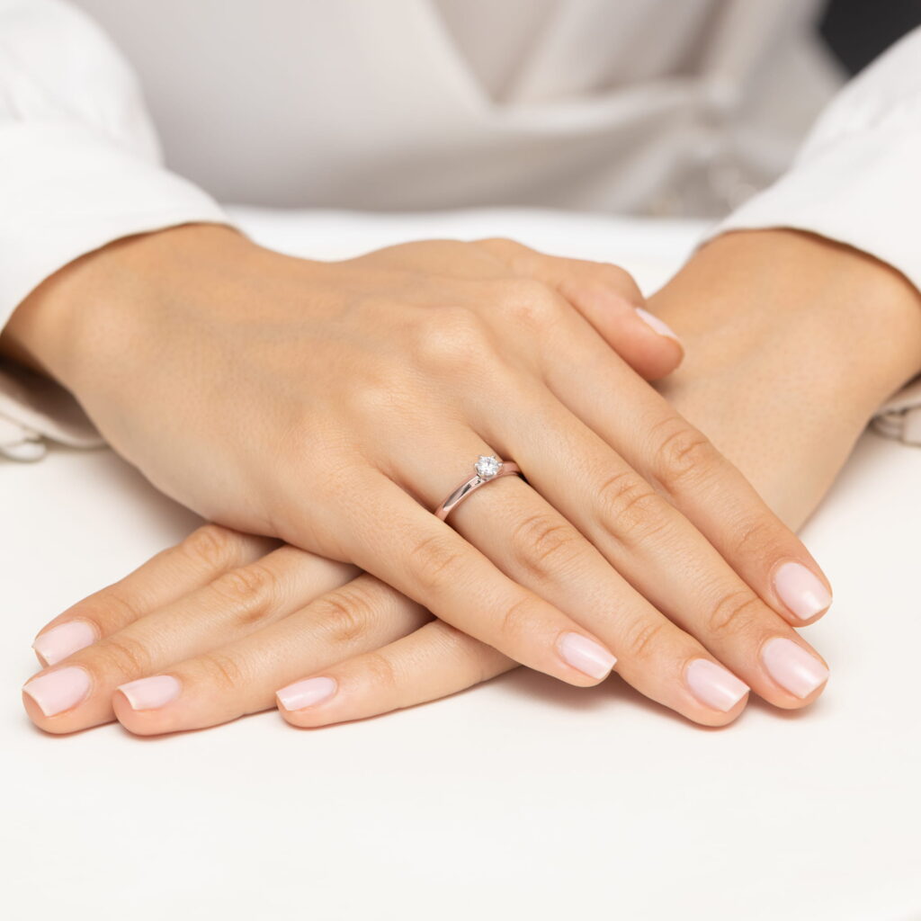 Dwie dłonie z delikatnym manicure, trzymające pierścionek z diamentem na palcu.