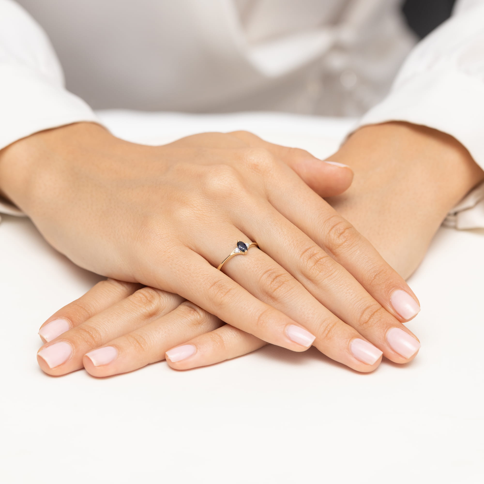 W0681.Z3.SAB.01_4 Dwie dłonie z delikatnym manicure, złożone na stole, z widocznym pierścionkiem z niebieskim kamieniem.