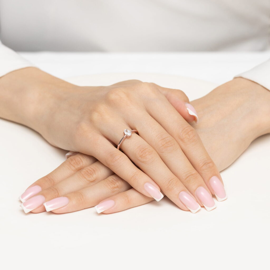 Dwie ręce z różowym manicure, jedna z pierścionkiem z diamentem, spoczywają na białym tle.