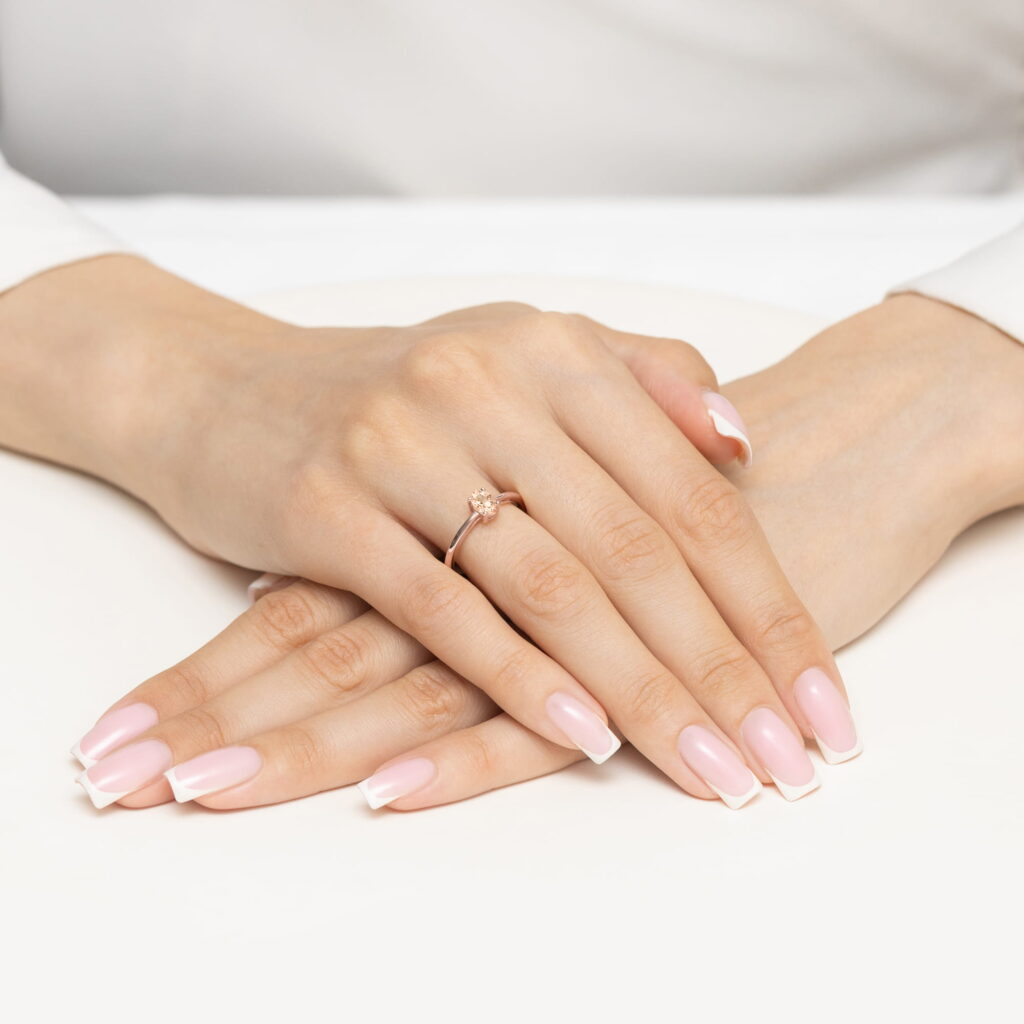 Dwie ręce z różowym manicure i pierścionkiem na palcu, ułożone na białym tle.
