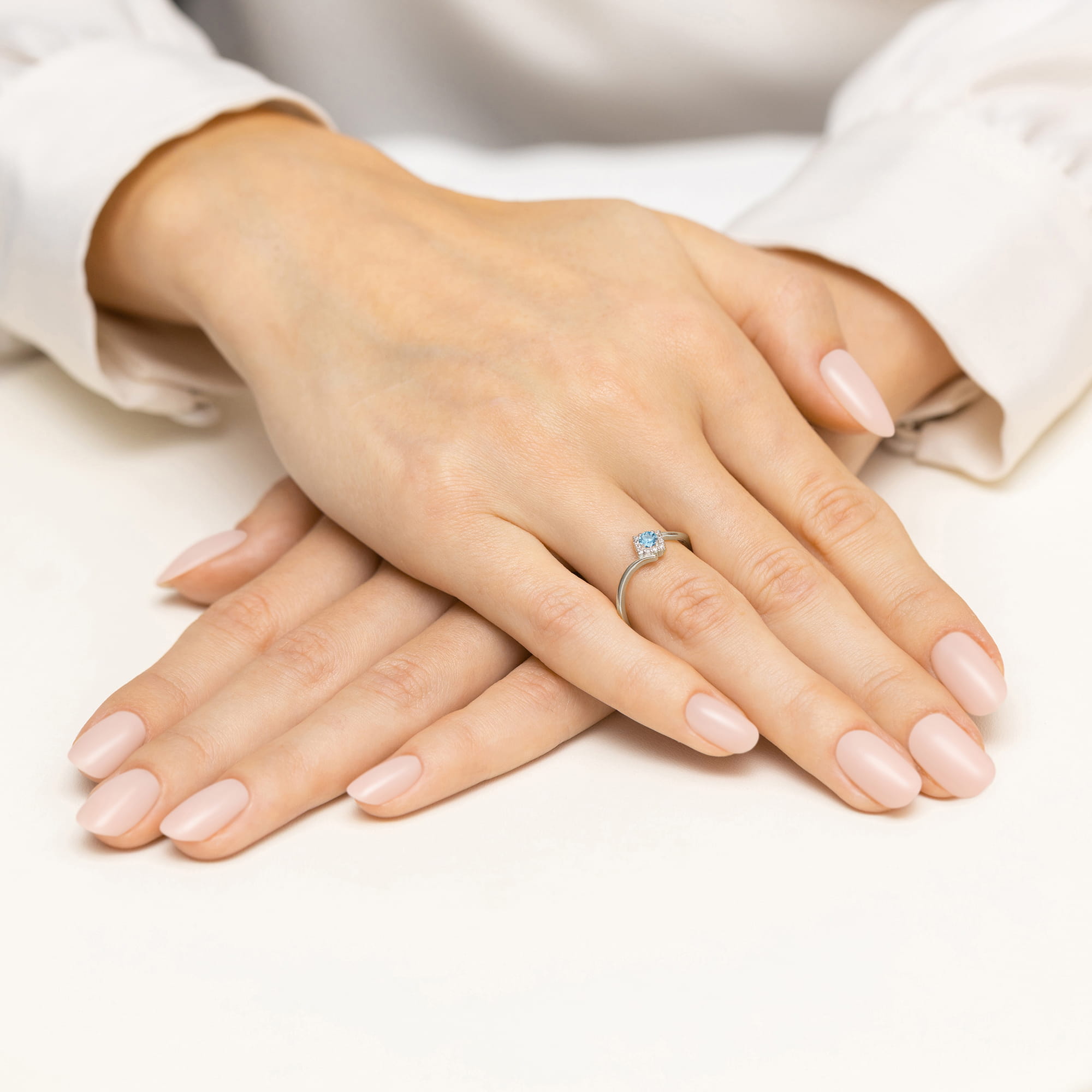 W0702.B3.TOB.01_4 Dwie ręce z delikatnym manicure, jedna z pierścionkiem z niebieskim kamieniem, ułożone na białym tle.