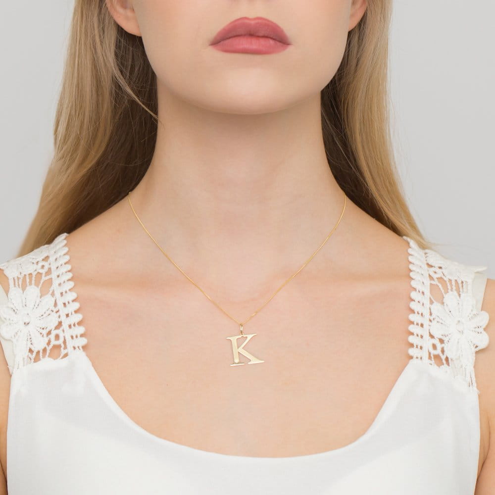 Z0070.Z3.DIW.11_3 Kobieta z długimi włosami nosi złoty naszyjnik z literą K na delikatnej, białej bluzce.